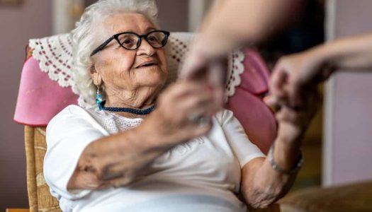 Female home carer supporting old woman to stand up from the armchair at care home